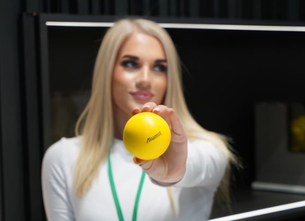 Person in white shirt holding a yellow stress ball with "Alumit" printed on it, blurred face, in an indoor setting.
